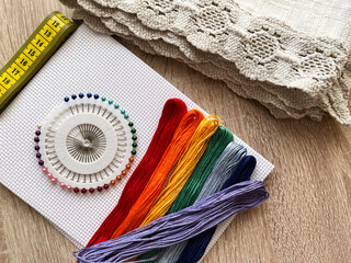 Multicolored floss threads, white canvas, sewing pins, and a centimeter ribbon lie on the table. Hobby or profession.