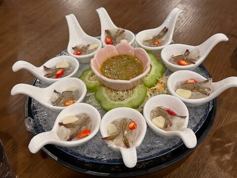 Thai Style Marinated Raw Shrimp. A platter featuring small dishes shaped like spoons, each containing shrimp, garlic, and chili, with spicy sauce in the center