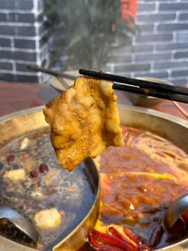 A piece of spicy lamb slice held with chopsticks over a bubbling hot pot filled with broth and spices.