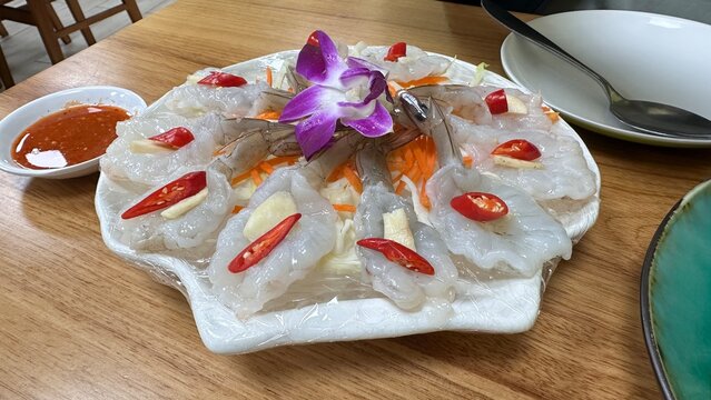 A plate of thai style marinated raw shrimp garnished with red chili slices and garlic, accompanied by a spicy dipping sauce on a wooden table.