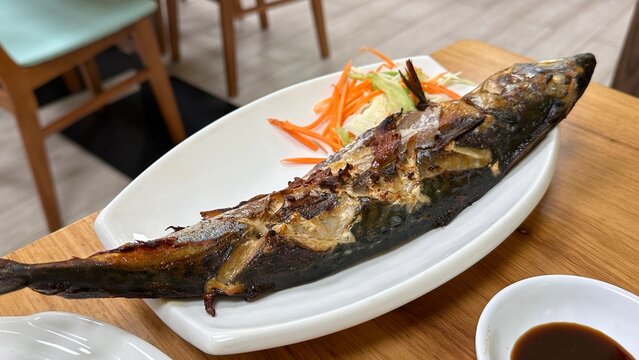 Thai style grilled Saba Mackerel served on a white plate, accompanied by shredded carrots and lettuce, with a small dish of sauce on the side