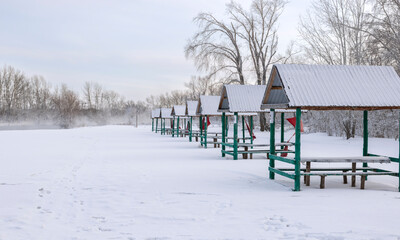 Snow-covered summer gazebos on the beach in winter near the forest
