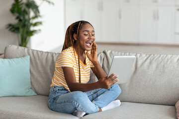 Obraz premium Happy stylish young black woman in casual student having video call with friends or family, sitting on couch in living room, using modern digital tablet, copy space. Telecommunication concept