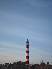 Panoramic view of Osinovetsky lighthouse with red and white stripes above pine forest. Iconic navigation tower against vast blue winter sky