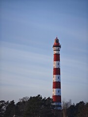 Tall red and white striped lighthouse standing behind winter forest trees against a clear blue sky. Osinovetsky lighthouse landmark at Lake Ladoga