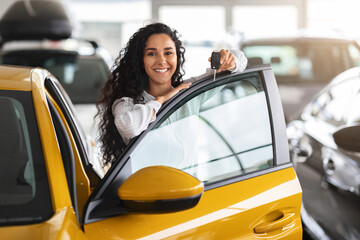 Cheerful brunette woman buying new cute car, raising hand up with automatic key and smiling, showroom interior, copy space. Pretty curly lady customer purchasing yellow female auto, getting in © Prostock-studio