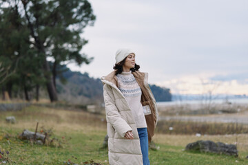Obraz premium Woman in a warm coat and hat enjoying outdoor scenery by a lake, embracing winter fashion amid natural landscape with trees and calm breeze