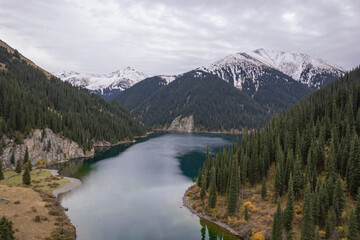 Aerial view of a calm mountain lake surrounded by evergreen forest and alpine slopes, with snow-capped peaks in the background and reflections on the water.
