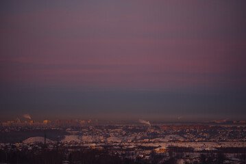 Naklejka premium Urban landscape features a vast expanse of snow-covered houses and buildings under a dramatic purple and blue sky during a cold winter sunset, highlighting the silhouette of the city horizon