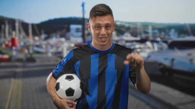 Man in blue striped jersey holding a soccer ball and winking with hand on hip at marina pier beside yachts and water; playful team spirit.