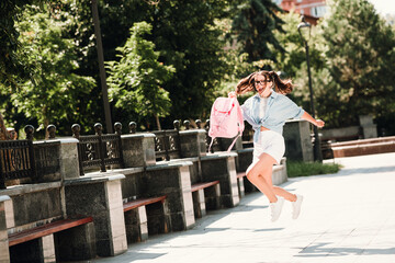 Obraz premium Young woman jumps with pink backpack in sunny park enjoying outdoor city life and casual fashion moment showcasing happiness energy youth friendship and active lifestyle in a bright urban street scene