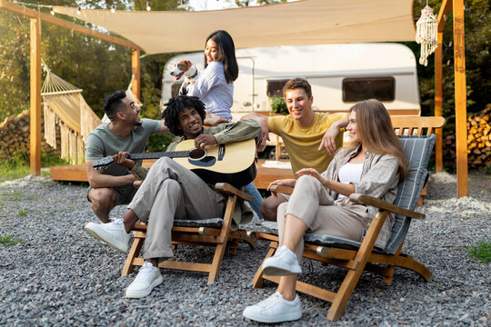 Young multiracial friends playing guitar and singing, holding pet dog, sitting in lounge chairs near RV, camping together, having fun autumn vacation, spending time in countryside