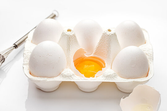 Close-up of a carton of six eggs with one cracked open on a table with a metal whisk