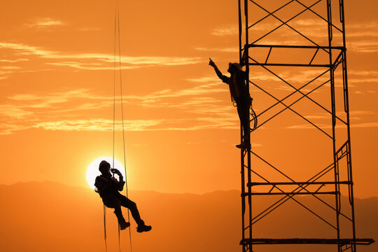 Silhouette of a construction worker hanging in a harness with another person standing on structural steelwork at sunset, Thailand