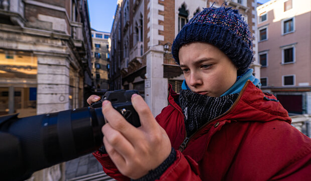 Close-up of a Teenage boy in a puffer coat and woolly hat reviewing photos on a digital single-lens reflex camera with a long lens, Venice, Veneto, Italy