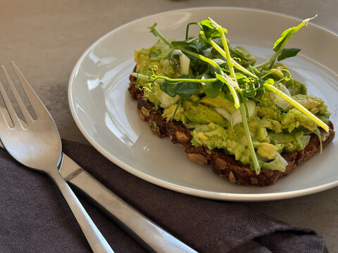 Close-up of a piece of toasted rye bread with avocado, spring onions and pea shoots on a plate with cutlery