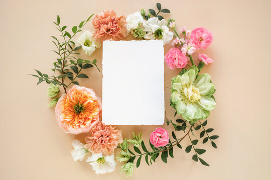 Close-up of a blank deckle edge card in the middle of a Floral wreath made of peony-shaped roses, lisianthus, carnation flowers and pistachio twigs on a beige background
