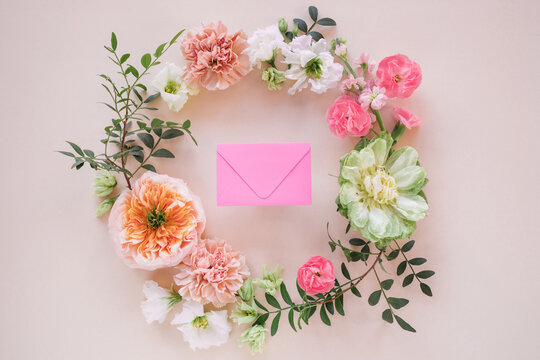 Close-up of a pink envelope in the middle of a Floral wreath made of peony-shaped roses, lisianthus, carnation flowers and pistachio twigs on a pink background