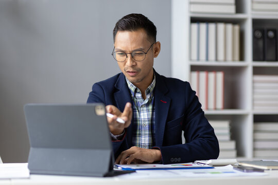 Stressed Asian businessman having video call on tablet in office, analyzing financial charts and graphs.