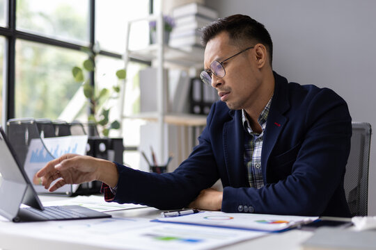Stressed Asian businessman having video call on tablet in office, analyzing financial charts and graphs.