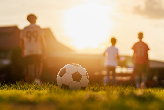 Soccer ball on green grass field with blurred silhouette of children playing football at sunset, golden hour light. Kids sport training concept.