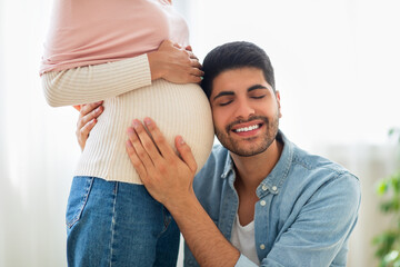 Delighted eastern man embracing tummy of his pregnant wife, listening baby kicks. Happy man...