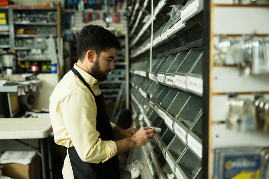Young man employee wearing apron sorting small parts from storage bins into a bag at hardware store