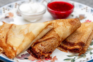 Traditional russian blini (thin pancakes or crepes) served folded with fresh strawberries, sour cream, and raspberry jam on a patterned plate