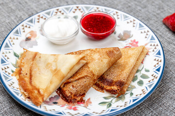 Traditional russian blini (thin pancakes or crepes) served folded with fresh strawberries, sour cream, and raspberry jam on a patterned plate