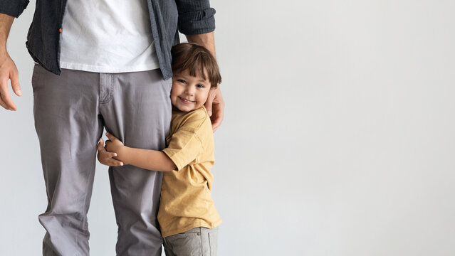 Father and son connection. Happy little boy embracing leg of his unrecognizable daddy, smiling to camera, feeling loved and safe, posing over grey wall background with empty space, panorama