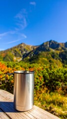 A silver mug sits on a wooden surface with a breathtaking mountainous backdrop, showcasing fall foliage and a bright blue sky