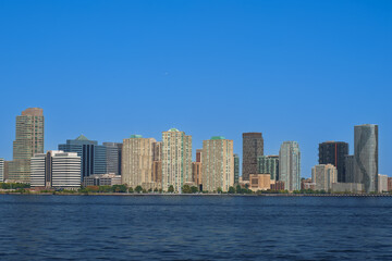 Obraz premium Panoramic view of the Jersey City skyline with modern glass skyscrapers reflecting sunlight under a clear blue sky, seen from the Hudson River.