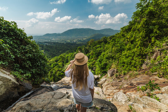 Young woman wearing straw hat standing on rocky ledge overlooking lush tropical valley and distant mountains near Samui waterfall in Thailand under bright sky and vibrant jungle scenery