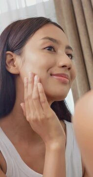 vertical clip. Beautiful Asian woman with tanned skin looking at mirror and applying skincare cream on her face