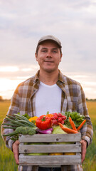 Farmer with a vegetable box in front of a sunset agricultural landscape. Man in a countryside field. Country life, food production, farming and country lifestyle concept.