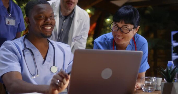 Multiethnic medical team sharing a genuine moment of laughter while reviewing patient data on a laptop, Japanese female doctor and African male colleague laughing during a clinical meeting.