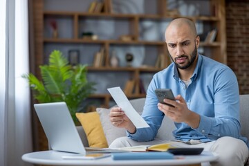 Man stressed at home table holding a bill and smartphone, laptop open as he calculates monthly...