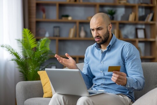 Frustrated man making an online purchase using a laptop at home, experiencing issues with his credit card transaction or payment, resulting in a declined payment
