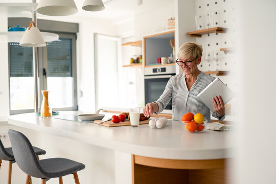 Cheerful senior woman enjoying bright morning working from home kitchen with tablet and breakfast