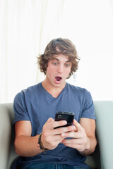 Man in his twenties sitting on light sofa holding black smartphone wearing blue t-shirt and jeans