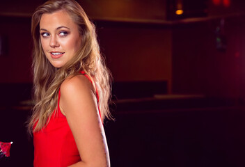 Naklejka premium Woman mid-20s turning, glancing over shoulder wearing sleeveless red dress in bar under warm lights