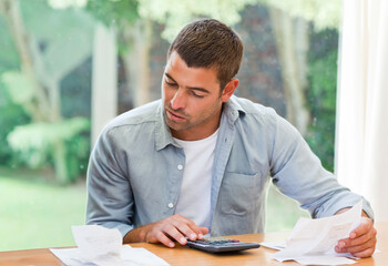 Mid-adult man wearing light blue shirt reviewing receipts while using calculator at wooden table