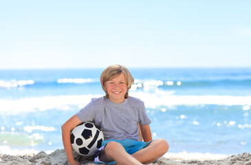 Child boy sitting cross-legged on sand holding black-and-white soccer ball at sunny beach