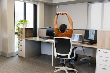 Dual monitors and laptop are displaying charts on wood-tone desk at open-plan office, with plant