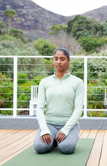 Obraz premium Indian woman kneeling on green yoga mat on wooden deck, eyes closed, in athletic wear