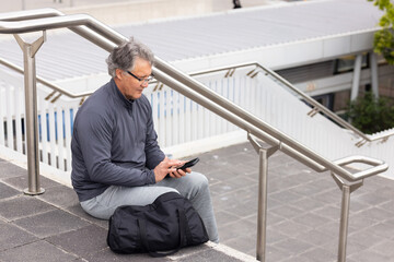 Obraz premium Senior man sitting on steps wearing gray sweats using smartphone, duffel bag nearby, copy space