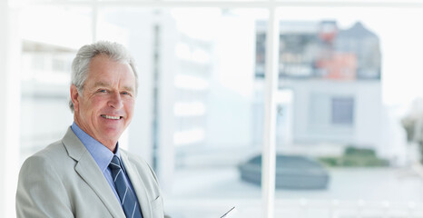 Senior man standing near glass windows in office in gray suit holding reading glasses, copy space