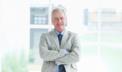 Senior man standing arms folded in bright lobby near windows wearing gray suit blue shirt tie