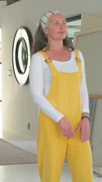 Vertical video: Entering woman in yellow overalls walking, inspecting canvases at home near mirror