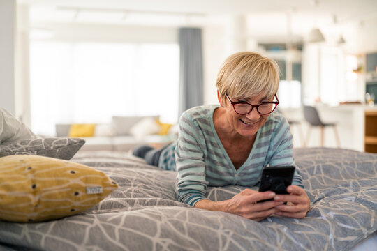 Bright natural light senior woman smiling using smartphone while lying on bed at home